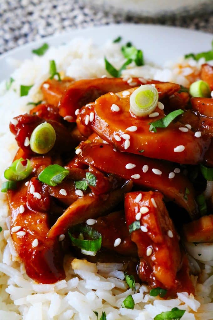 A close up shot of sliced teriyaki chicken over white rice garnished with sesame seeds and green onions.