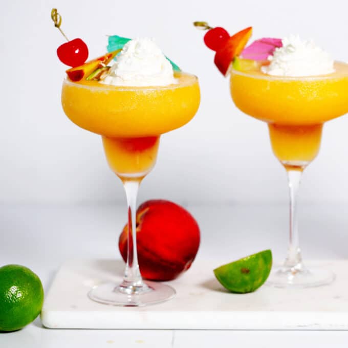 Two peach daiquiris in margarita glasses topped with fruited skewer, whipped cream, and a cocktail umbrella. They are on a white marble tray with peaches and limes around them.