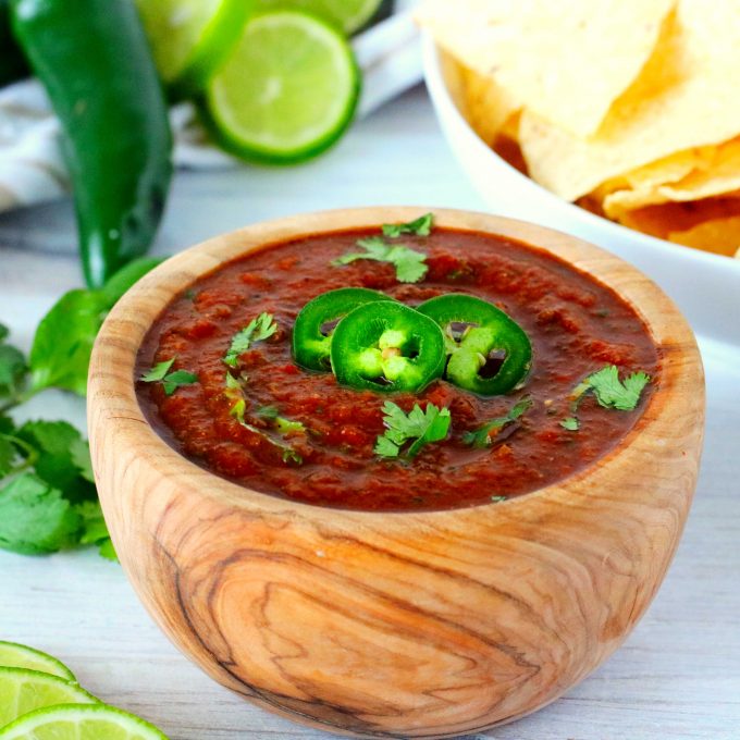 A small wood bowl with Mexican salsa inside. The bowl is surrounded by peppers, limes, and cilantro.