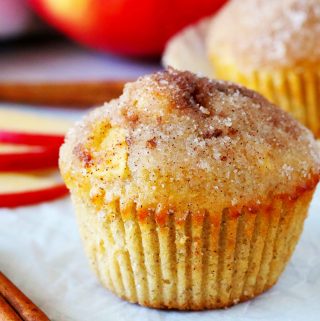 An apple cinnamon muffin with a cinnamon stick beside it and sliced apples in the background.