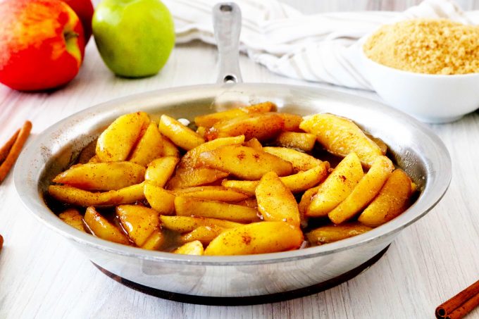 Fried apples in a pan with a bowl of brown sugar and apples behind it. 