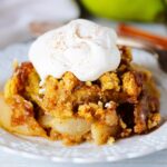 Pear cobbler topped with whipped cream and cinnamon on a white plate with a fork and pears behind it.