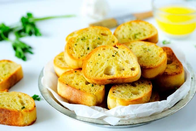 Garlic bread toast in a silver dish with a white napkin liner. There is parsley and garlic butter in the background.