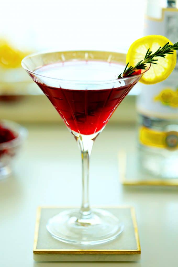 A close up shot of a pomegranate martini with a lemon wheel wedge and rosemary garnish.