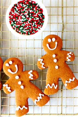 Two gingerbread cookies on a cooling rack with a bowl of sprinkles off to the side.