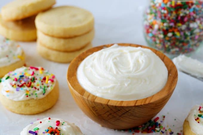 A bowl of white sugar cookie frosting with cookies and sprinkles in the background.