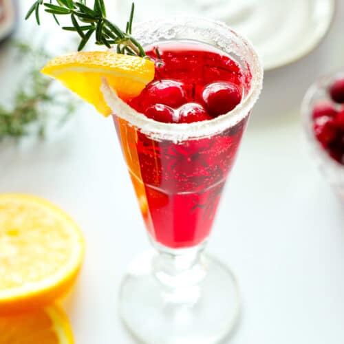 A cranberry mimosa with a sugar rim and an orange, rosemary, and cranberry garnish with a plate of sugar and a bowl of cranberries in the background. There are also some orange wedges off to the side.