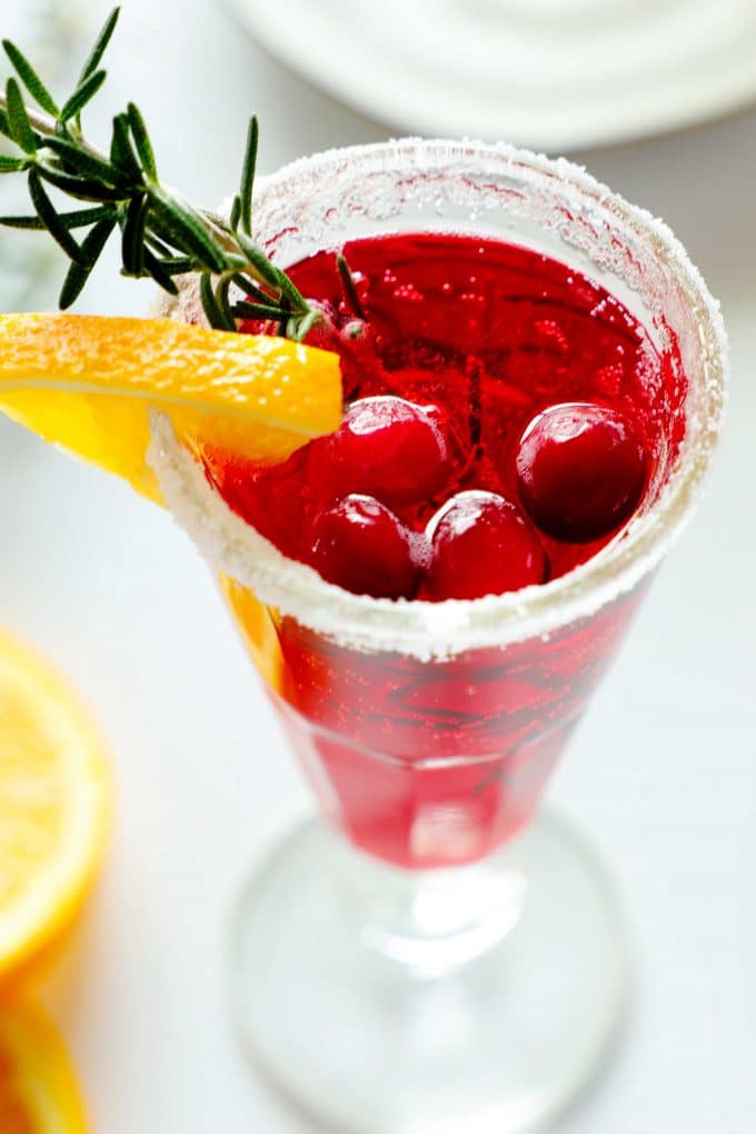 A close up shot of A cranberry mimosa with a sugar rim and an orange, rosemary, and cranberry garnish.