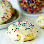 Sour Cream Cookie on a white surface with a plate of cookies and a bowl of sprinkles behind it.