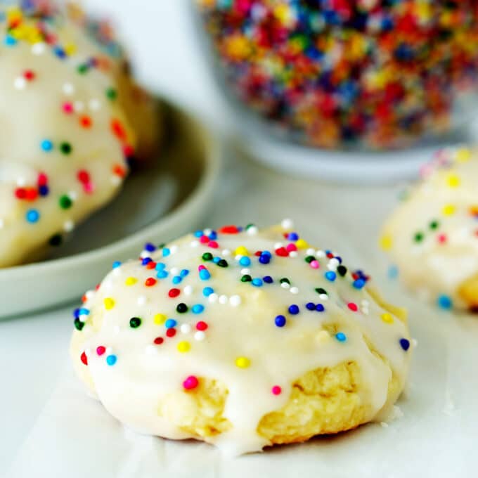 Sour Cream Cookie on a white surface with a plate of cookies and a bowl of sprinkles behind it.