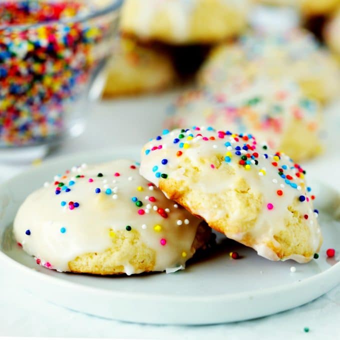 Two sour cream cookies on a white plate with cookies and sprinkles in the background.