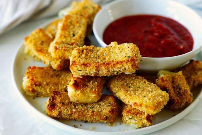 Air fryer mozzarella sticks on white plate with a bowl of marinara sauce off to the side. 