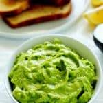 Avocado spread in a bowl with toast off to the side and lemon wedges.