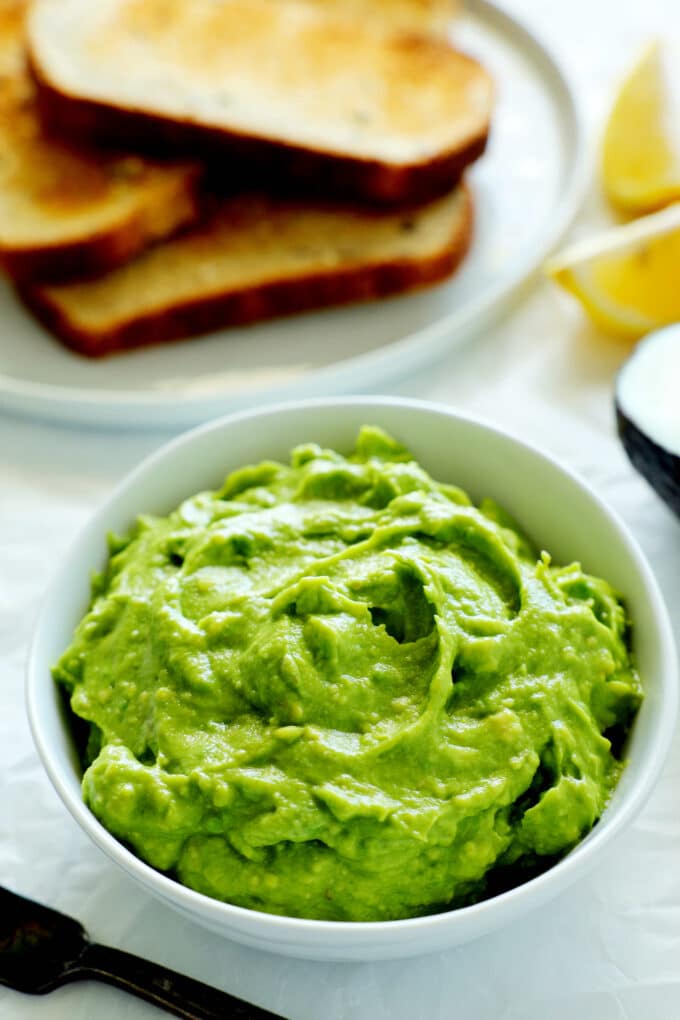 Avocado spread in a bowl with toast off to the side and lemon wedges. 