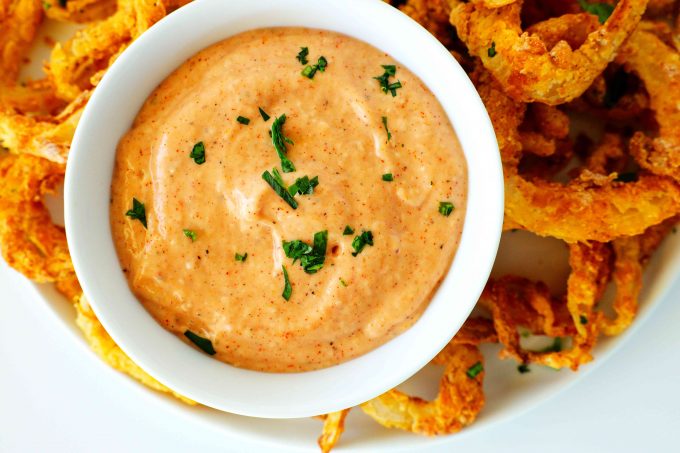 A close up shot of comeback sauce in a white bowl on a plate with Air Fryer Onion Rings.
