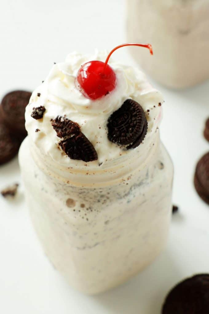 A close up shot of a cookies and cream shake topped with oreos, whipped cream, and a cherry. There are oreos and another shake in the background.
