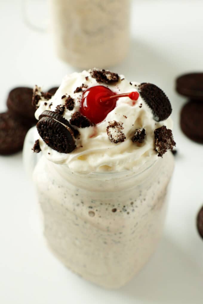 Cookies and cream shake topped with oreos, whipped cream, and a cherry. There are oreos and another shake in the background.