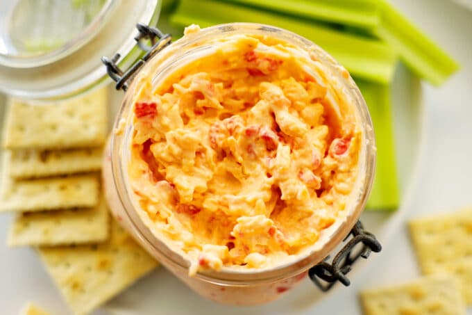 An overhead shot of pimento cheese in a jar on a plate with celery and crackers. 