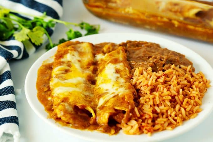 Two cheese enchiladas with enchilada gravy on a plate with rice and refried beans. There is a striped towel and cilantro sprigs off to the side.