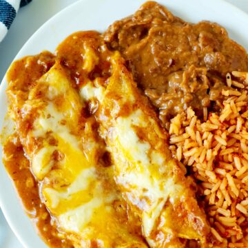 An overhead shot of two cheese enchiladas with enchilada gravy on a plate with rice and refried beans.