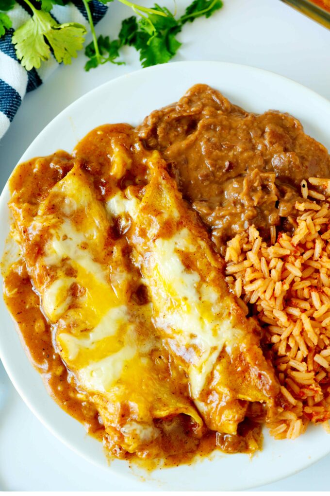 An overhead shot of two cheese enchiladas with enchilada gravy on a plate with rice and refried beans.