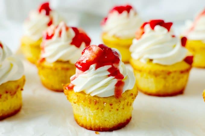 A group of strawberry shortcake cupcakes on a white surface all topped with whipped cream and fresh berries.