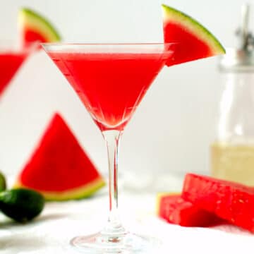 Two watermelon martinis on a white surface with limes and watermelon wedges surrounding them.