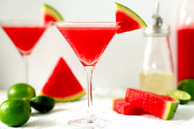 Two watermelon martinis on a white surface with limes and watermelon wedges surrounding them. 
