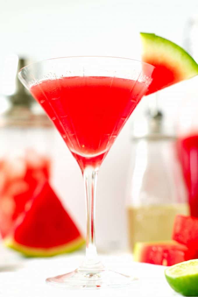 A close up shot of a watermelon martini with simple syrup and a shaker in the background.