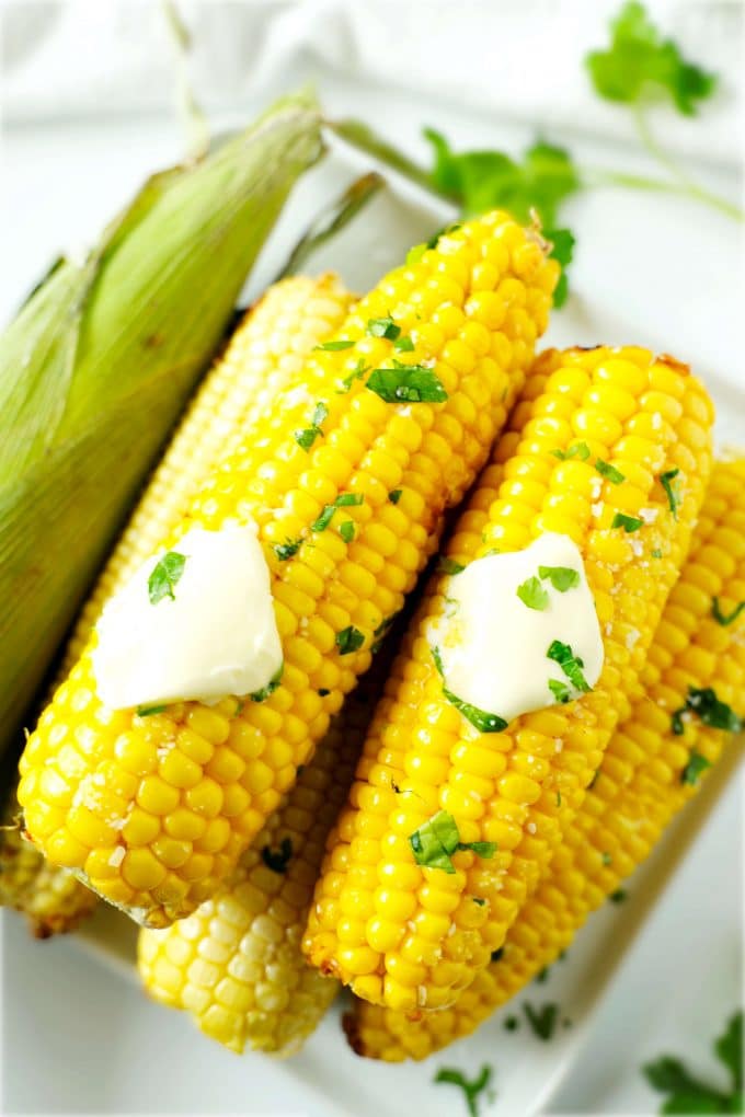 Air Fryer Corn On the Cob with butter pads on top of them stacked on a plate with a uncooked ear of corn off to the side.