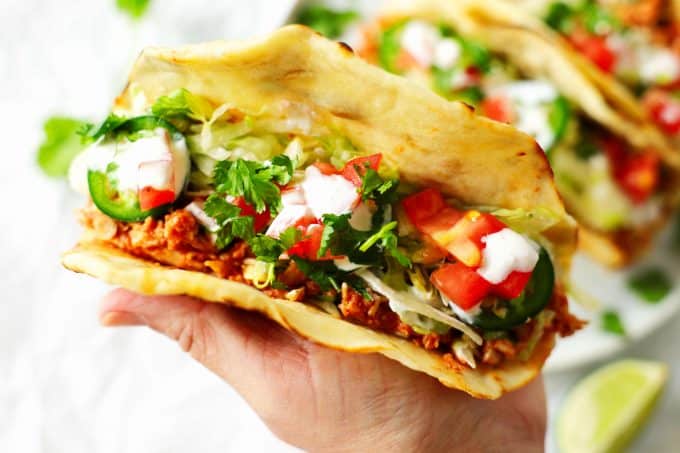 A hand holding a shredded chicken taco. The tacos are topped with sliced jalapenos, sour cream, pico de gallo, and cilantro.