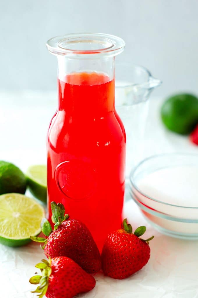 A bottle of simple syrup with strawberries, sugar, and limes off to the side.