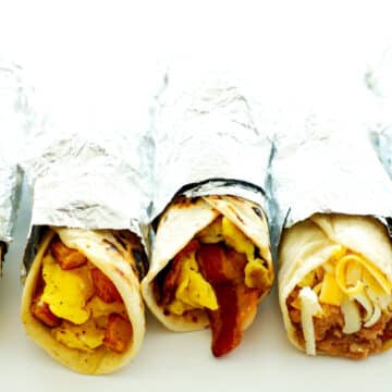Five different breakfast tacos wrapped in a aluminum foil on a white background.