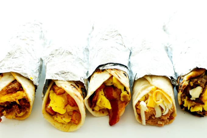 Five different breakfast tacos wrapped in a aluminum foil on a white background.