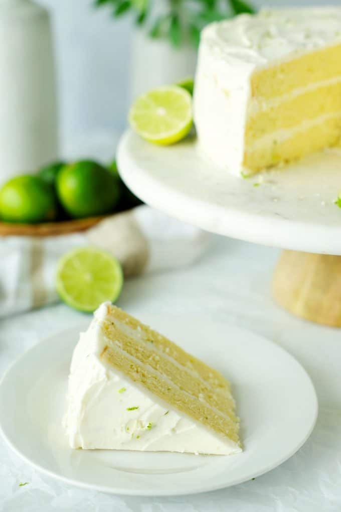 A slice of key lime cake on a plate with a cake on a cake stand behind it.