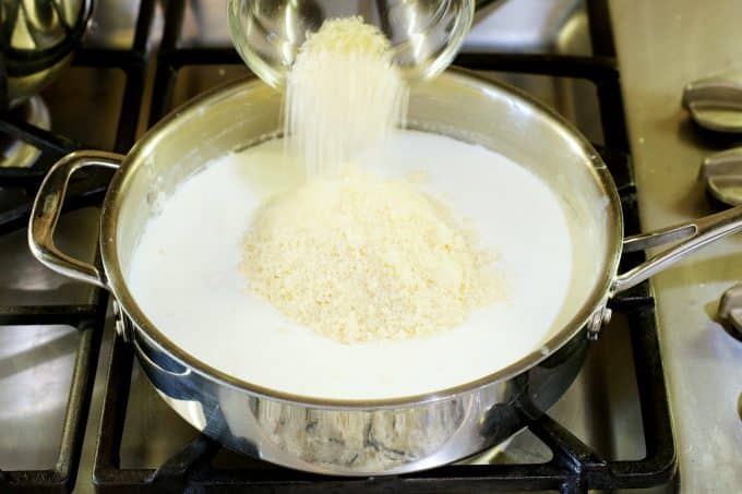 Parmesan being poured into a milk mixture in a sauce pan on the stove top.