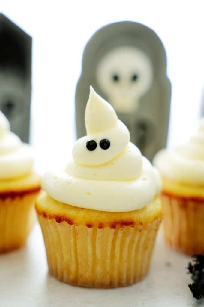 A vanilla ghost cupcake on a white surface with a grave behind it.