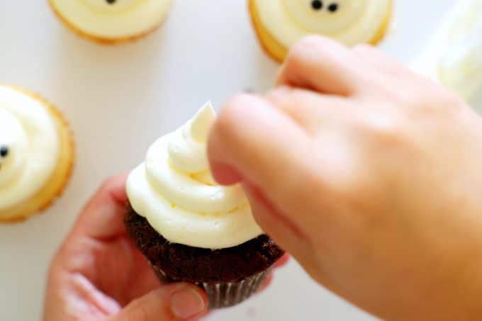 A hand putting black pearl sprinkles onto a swirl of white frosting on top of a chocolate cupcake.