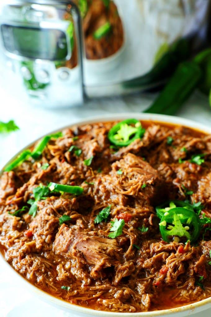 Mexican shredded beef in a white bowl in front of a slow cooker.