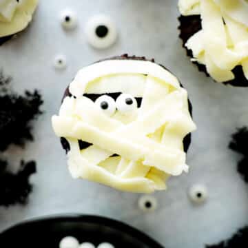 A white mummy cupcake on a white surface with black moss around it.