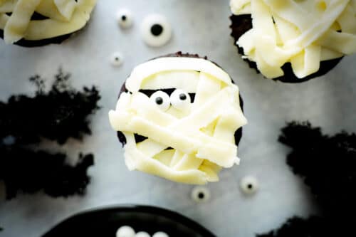 A white mummy cupcake on a white surface with black moss around it.