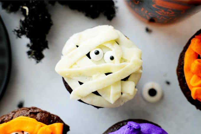 A mummy cupcake with white frosting and candy I decals around it.