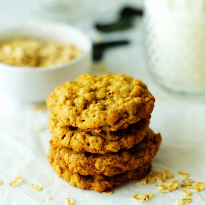 Old-fashioned oatmeal cookies stacked on a crinkled white surface with a glass of milk and oats behind them.