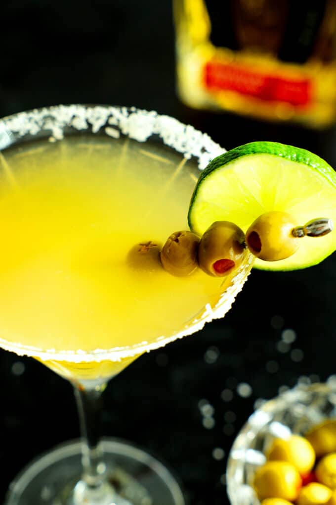 An overhead shot of a Mexican martini with a salted rim and a garnish of olives and limes.