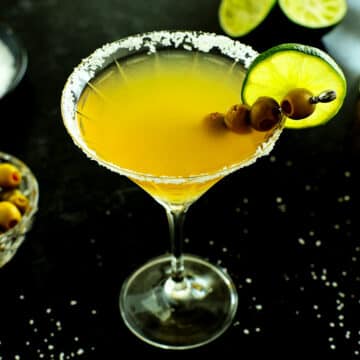 A Mexican martini on a black surface with olives around it and salt on a plate behind it.