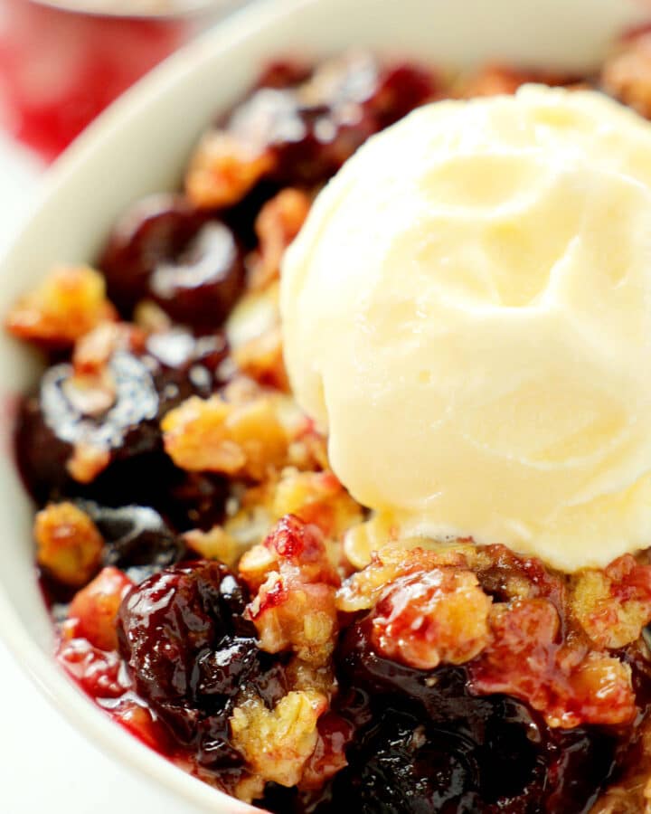 A close up of a bowl of cherry crisp with a scoop of vanilla ice cream