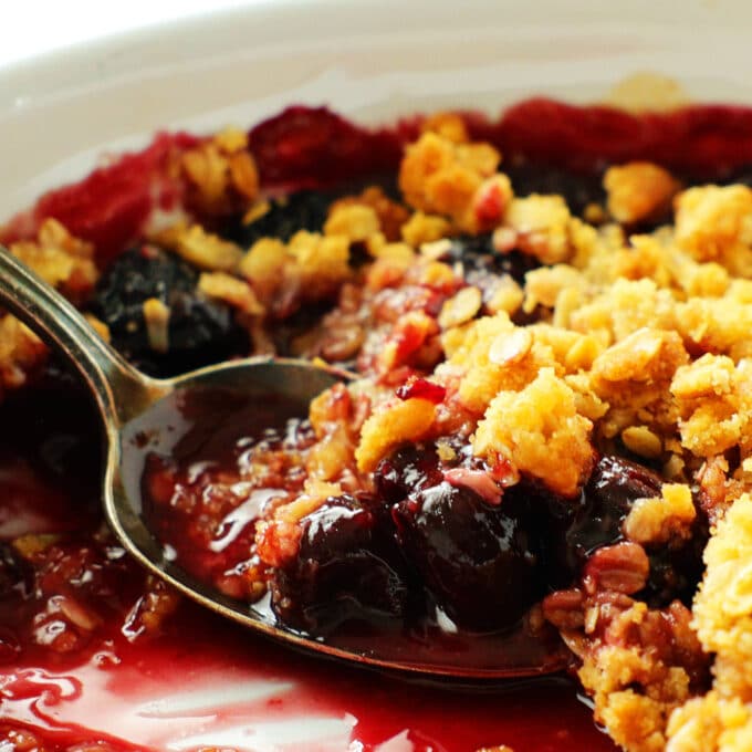 A close up of a spoon in a dish of cherry crisp with a scoop taken out of if