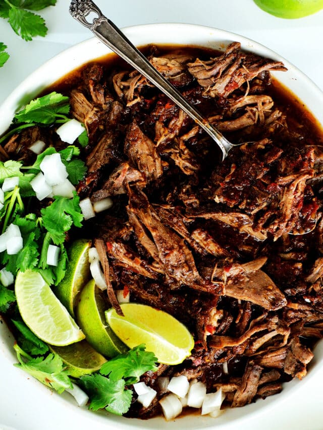 a close up of A bowl of beef birria stew with cilantro, lime, and onions on top