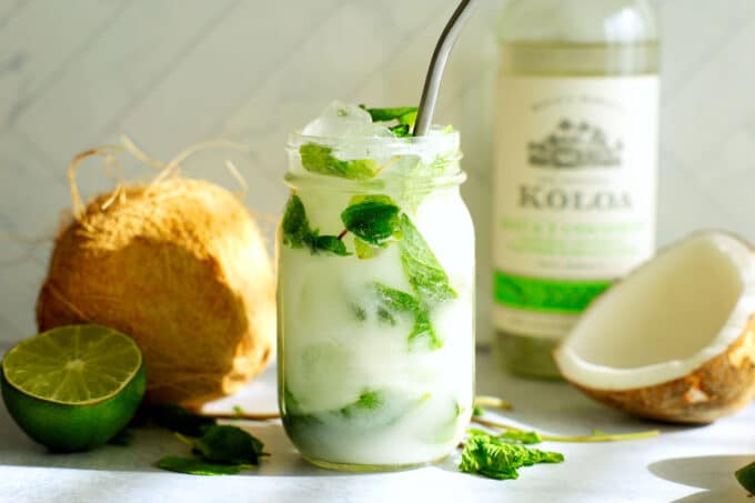 A coconut mojito in a jar with a straw. A coconut and limes in the background