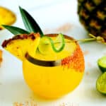 A pineapple margarita with a lime twist, pineapple wedge, and pineapple leaves garnish and tajin on one side of the glass